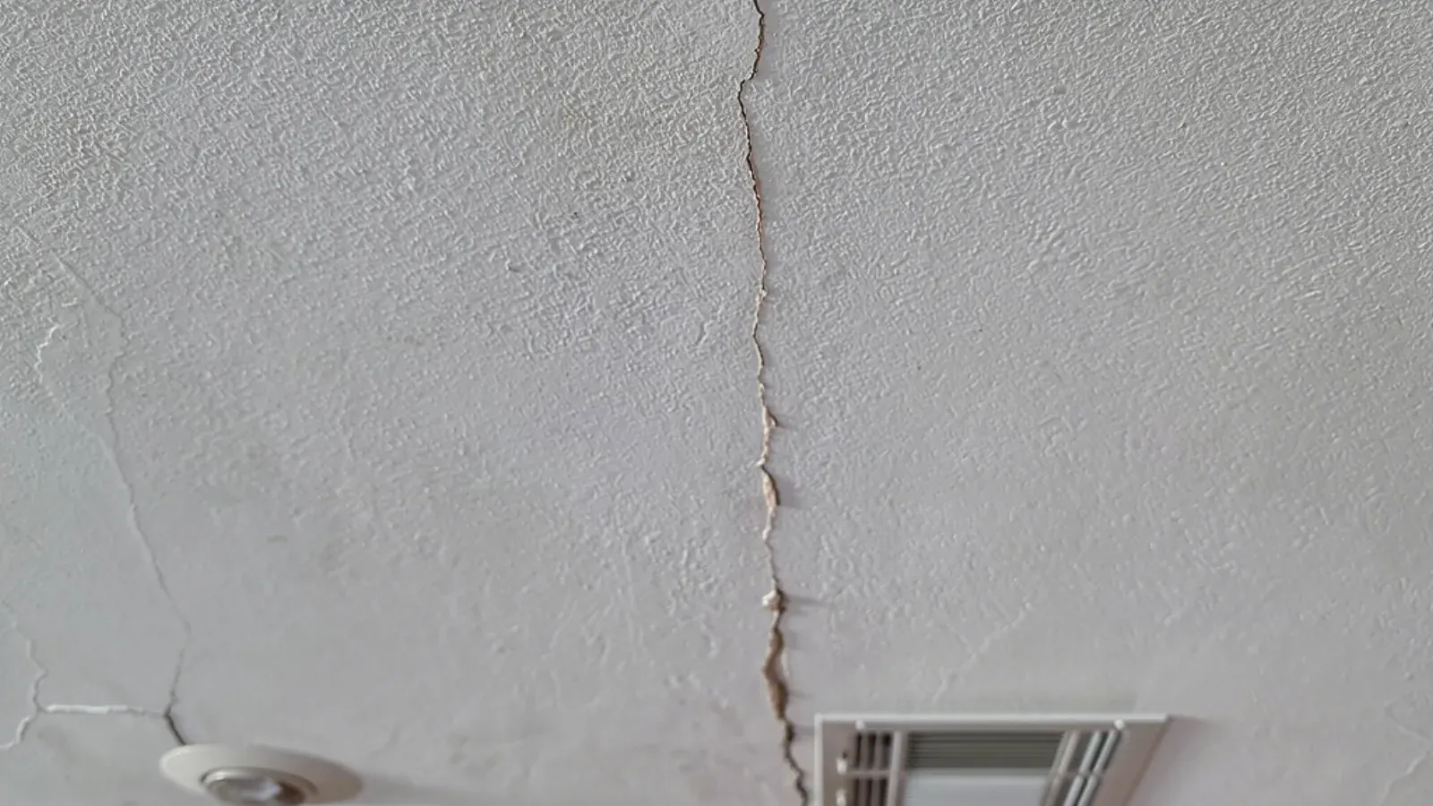 A crack split open along a joint in a textured painted ceiling, running past a round recessed light and a rectangular ceiling air register