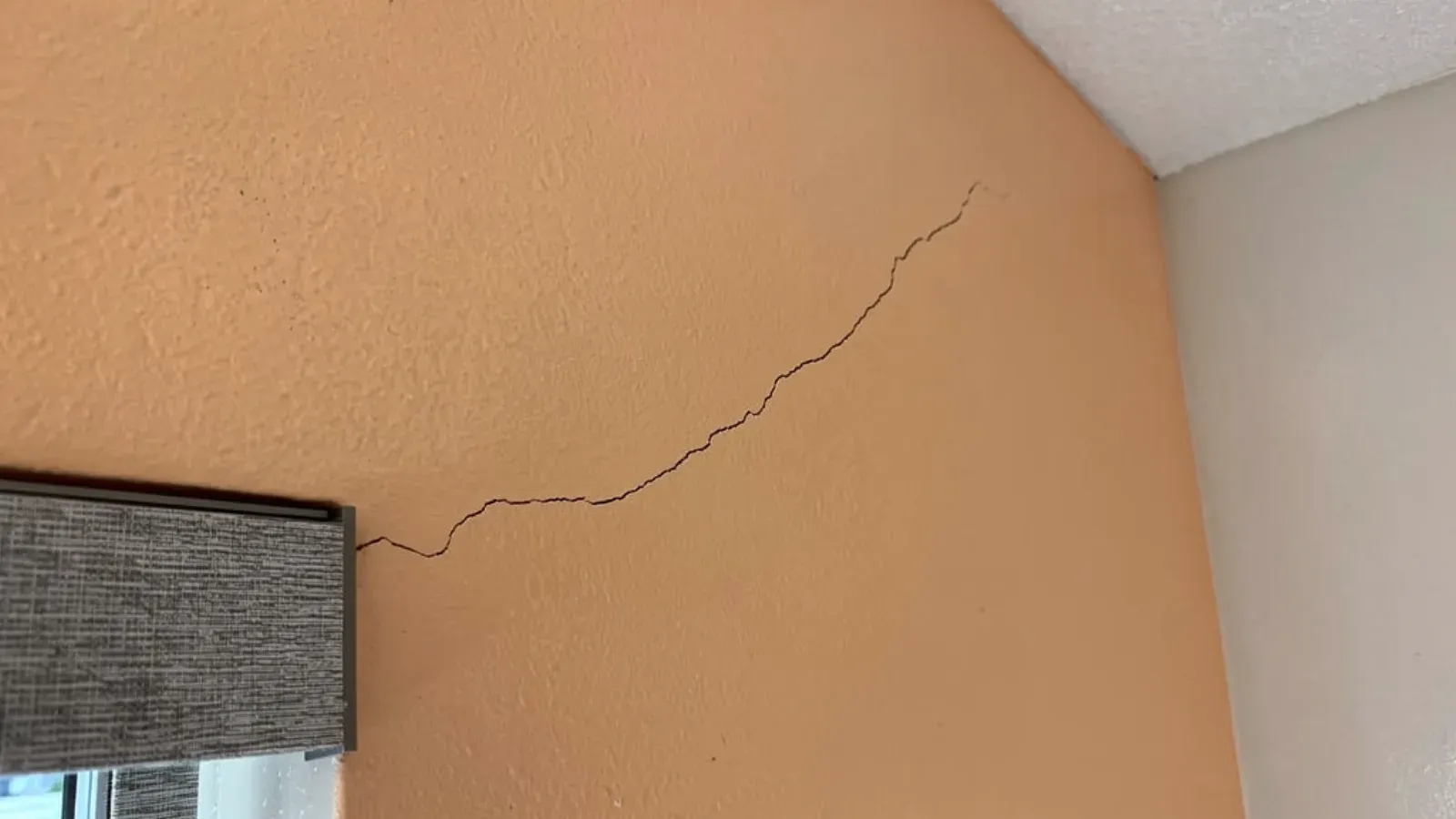 A hairline stress crack running diagonally across a painted interior wall from the head of a window casing up toward the wall-and-ceiling corner