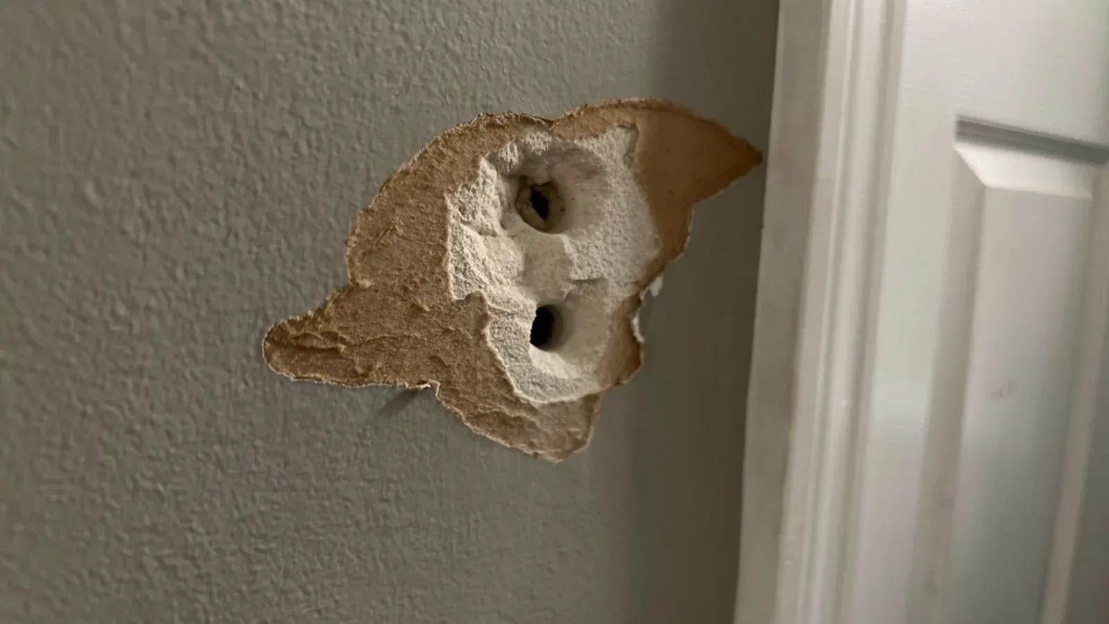 Impact hole in an interior drywall wall beside a door casing, torn paper facing peeled back exposing the gypsum core
