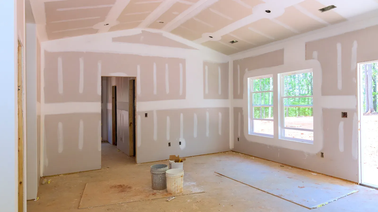 Newly hung drywall room with every seam taped and coated in white joint compound and each fastener spotted, still unsanded, with joint-compound buckets on the bare subfloor