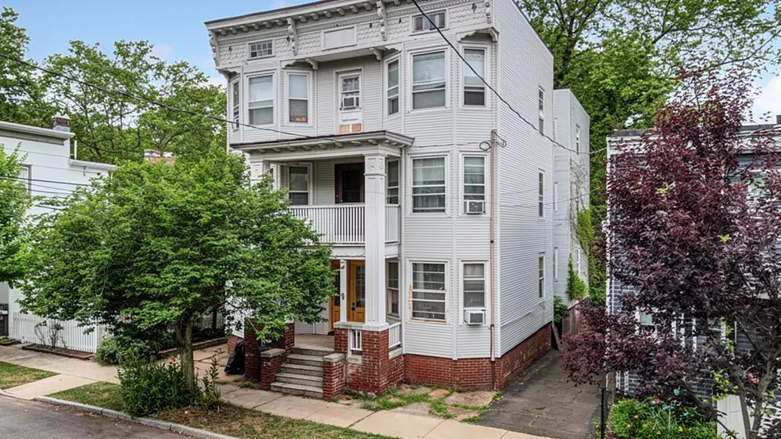 A three-story multi-family house with white siding, columned front porches and a brick foundation