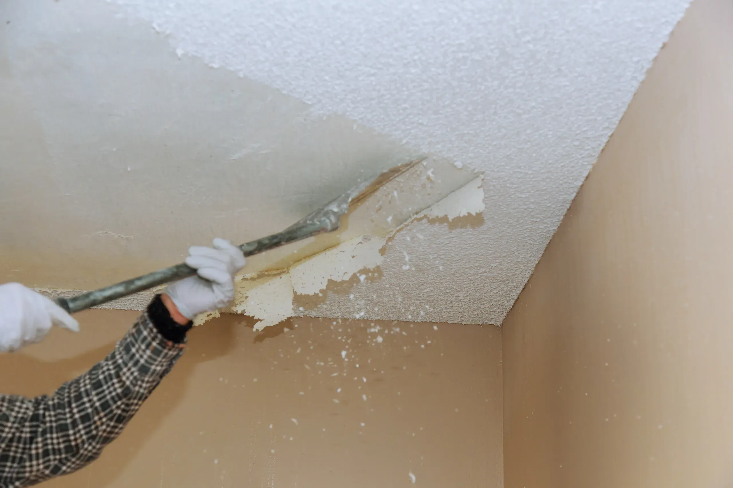 Long-handled scraper pushing along a popcorn ceiling, a wide band already stripped to bare board with the untouched texture beyond it